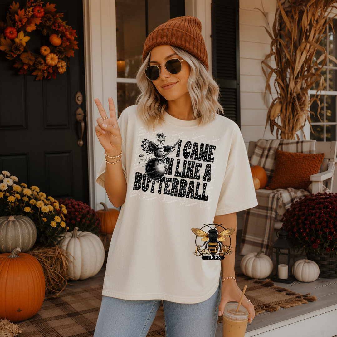 A woman stands on a decorated autumn front porch wearing an oversized cream graphic T-shirt with black text reading 'I CAME IN LIKE A BUTTERBALL' and a bee logo. She is also wearing a brown beanie, sunglasses, and light blue jeans, holding an iced coffee.