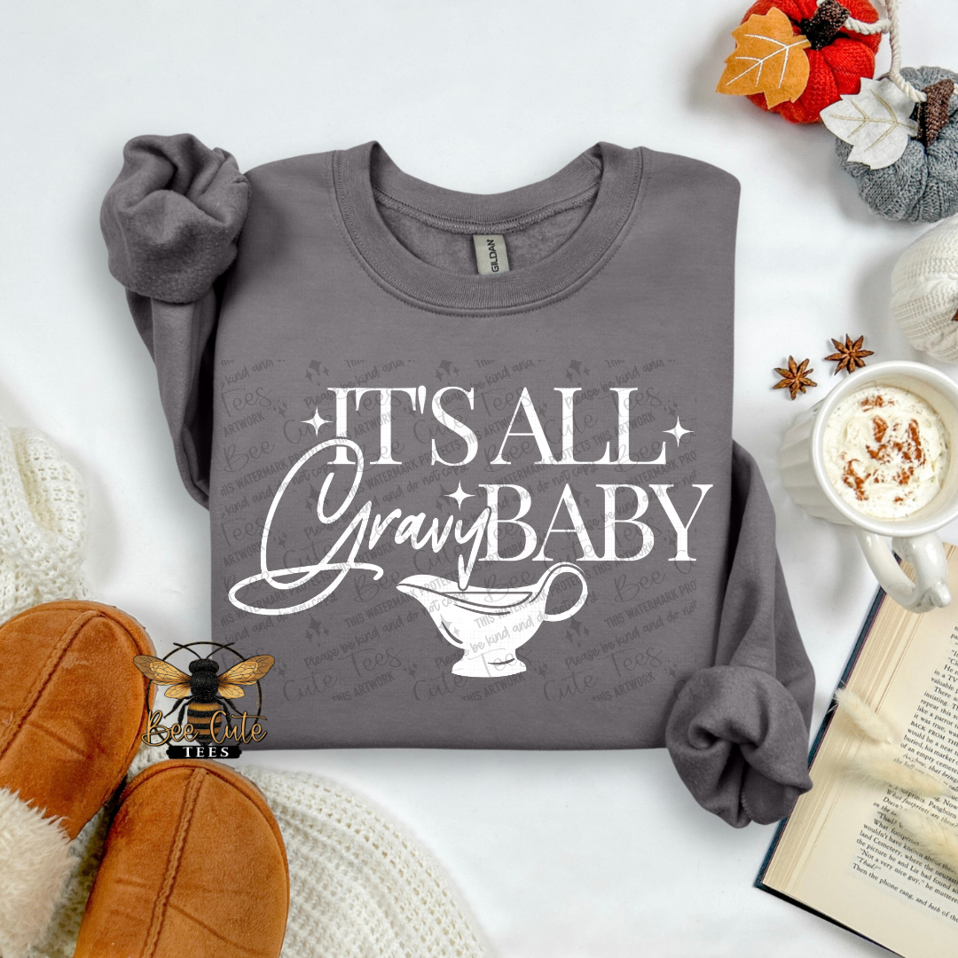 A folded dark gray sweatshirt with rolled sleeves, featuring the white text 'IT'S ALL GRAVY BABY' and a gravy boat graphic on the front. The sweatshirt is styled in a cozy flat-lay arrangement with autumn-themed props including a mug of hot chocolate, an open book, a small bee graphic, and fall decorations on a white background.