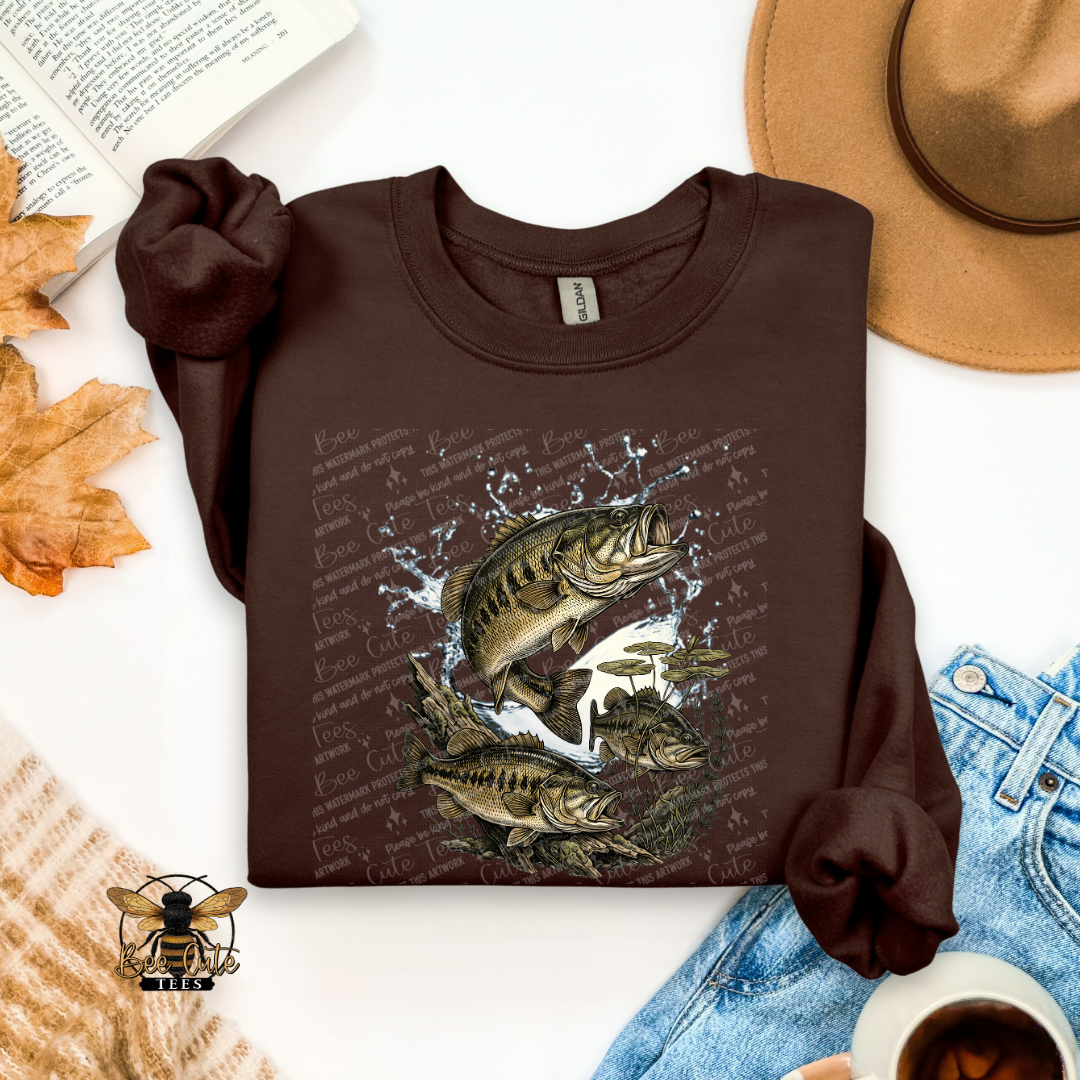 A dark brown long-sleeve t-shirt with a graphic of jumping bass fish and water splashes, styled in a flat lay with a tan hat, blue jeans, a book, autumn leaves, a cup of coffee, and a small bee logo.