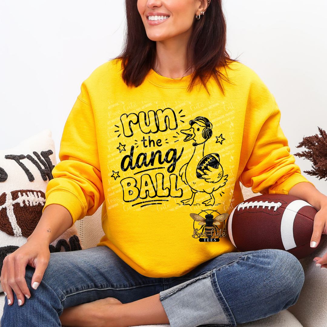A woman wearing a bright yellow sweatshirt with a black graphic that reads 'Run the dang ball' and a duck illustration, sitting cross-legged and holding a football.