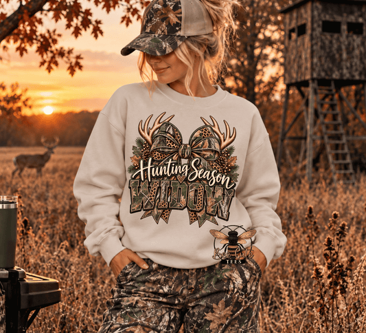 A woman wearing a camouflage hunting cap and camouflage pants stands in a field at sunset, wearing a cream sweatshirt with a large 'Hunting Season Widow' deer graphic on the front.