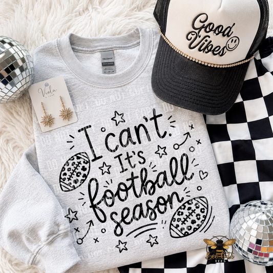 A light gray crewneck sweatshirt with a black graphic reading 'I can't. It's football season' with football and star doodles, styled in a flat lay with a black and white checkered cloth, a black and white trucker hat reading 'Good Vibe', gold star earrings, and two disco balls.
