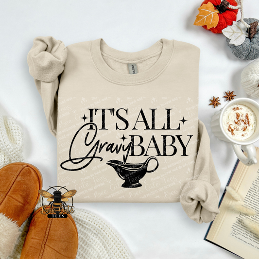 A beige long-sleeve crewneck sweatshirt with 'IT'S ALL Gravy BABY' and a gravy boat graphic printed on the front, styled in an autumn flat-lay with brown slippers, a mug of hot chocolate, an open book, and fall leaves.