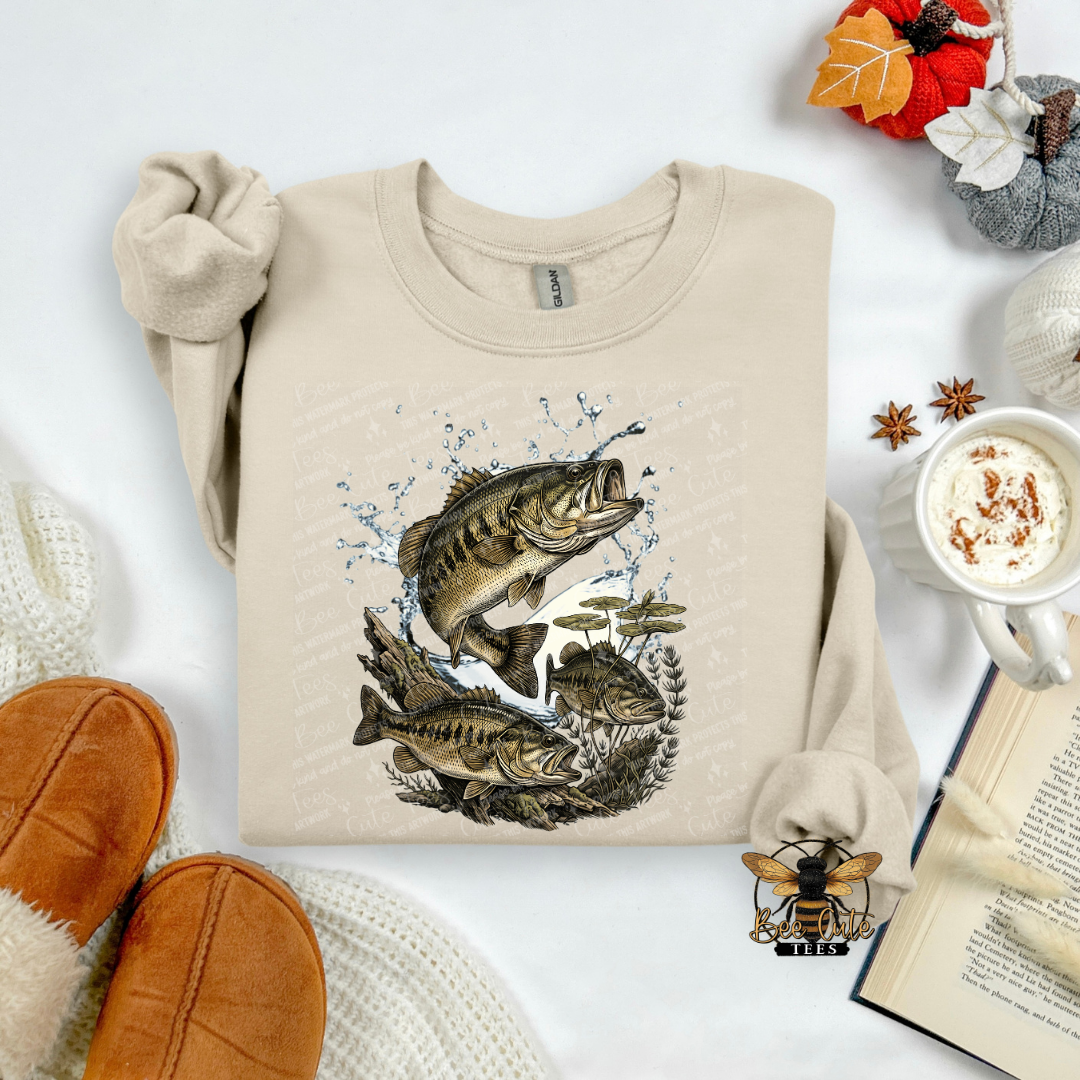 A beige crewneck sweatshirt with a graphic of three largemouth bass fish jumping out of water, styled in a cozy flat-lay arrangement with brown slippers, a mug of coffee, an open book, and autumn decorations.