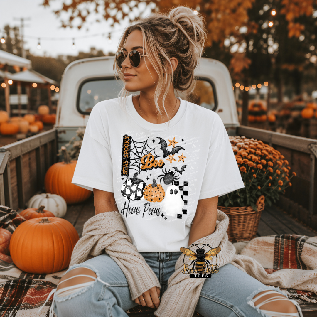 A woman wearing a white graphic t-shirt with a Halloween design sits in the bed of a pickup truck surrounded by pumpkins and fall decorations. The t-shirt features a graphic of a spider, moon, acorns, and the text 'Hocus Pocus'.