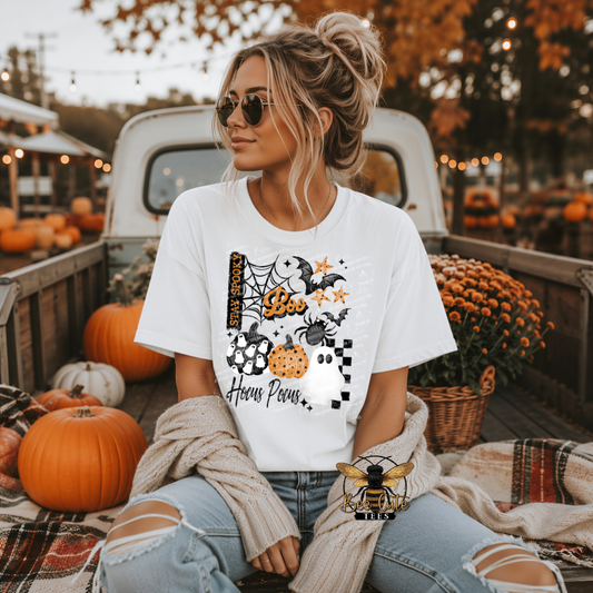 A woman wearing a white graphic t-shirt with a Halloween design sits in the bed of a pickup truck surrounded by pumpkins and fall decorations. The t-shirt features a graphic of a spider, moon, acorns, and the text 'Hocus Pocus'.