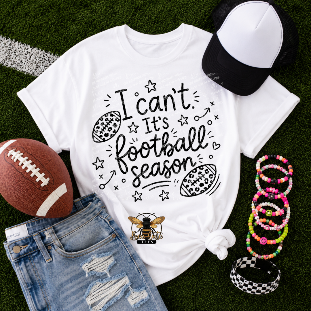 A white graphic T-shirt laid flat on green turf with a football, jeans, a black-and-white cap, and colorful beaded bracelets around it. The shirt has black text reading 'I can't It's football season' with football-themed doodles and a bee logo near the hem.