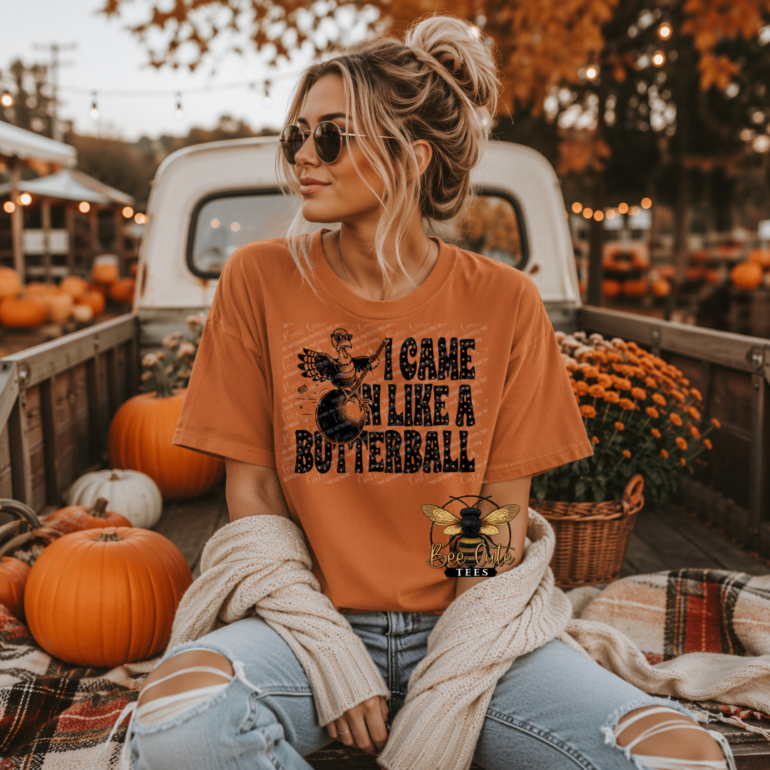 A woman wearing a burnt orange graphic t-shirt sits in the bed of a pickup truck surrounded by pumpkins and fall decorations. The t-shirt features a black graphic of a turkey and the text 'I CAME IN LIKE A BUTTERBALL'.