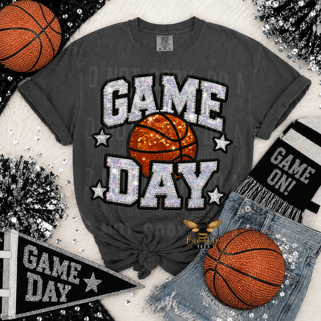 A dark gray graphic t-shirt with a 'GAME DAY' design featuring a basketball and white glitter-style lettering, styled in a flat lay with basketballs, denim shorts, a black knit beanie, and a black pennant banner on a white faux fur background.