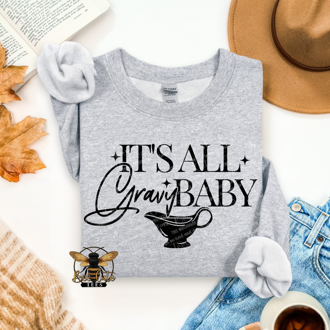 A folded heather gray graphic T-shirt with rolled sleeves, printed with the phrase 'IT'S ALL Gravy BABY' and a gravy boat illustration. The shirt is styled in a flat lay with an open book, autumn leaves, a tan hat, blue jeans, a knit sweater, and a small bee logo near the bottom.