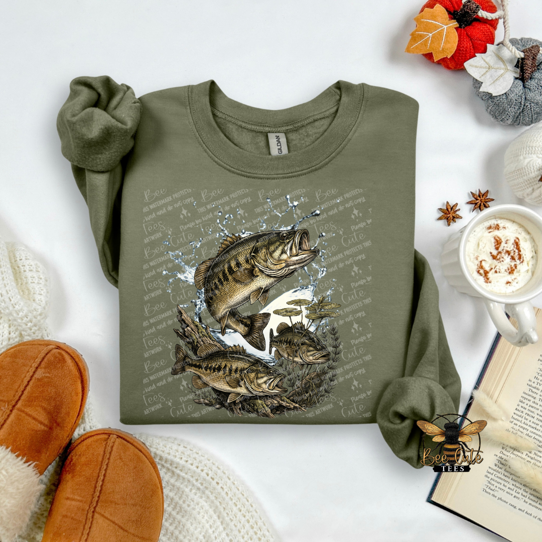 An olive green long-sleeve t-shirt with a graphic of three largemouth bass fish jumping through water, styled in a cozy flat-lay arrangement with a mug of coffee, an open book, and brown slippers.