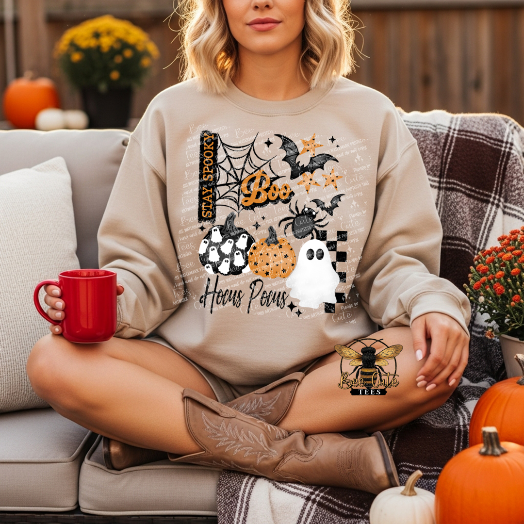A woman sits cross-legged on a couch wearing a beige crewneck sweatshirt with a colorful Halloween graphic that reads 'Boo Hocus Pocus' with pumpkins, ghosts, spiders, and bats. She holds a red mug in a cozy fall setting with pumpkins and a plaid blanket.