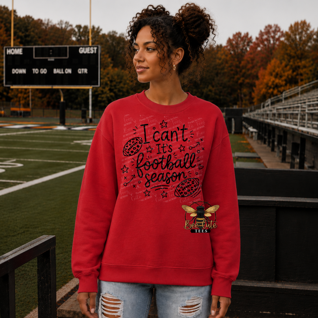 A woman stands on a football field wearing a red crewneck sweatshirt with a black graphic that reads 'I can't, it's football season' and a bee logo underneath. The background shows a football field, stadium seating, and trees with autumn foliage.