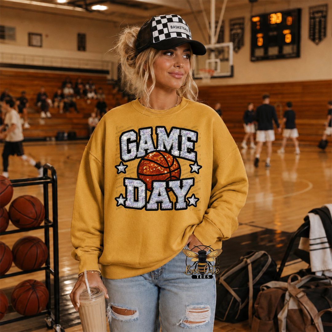 A woman stands on a basketball court wearing a mustard yellow sweatshirt with a 'GAME DAY' graphic featuring a basketball and stars, paired with a black and white checkered cap and ripped light-wash jeans.