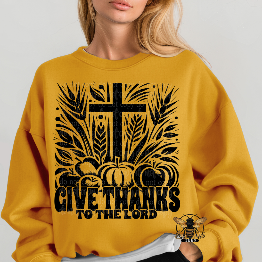 A woman wearing a mustard yellow sweatshirt with a black graphic print featuring a cross, pumpkins, wheat, and the text 'GIVE THANKS TO THE LORD'.