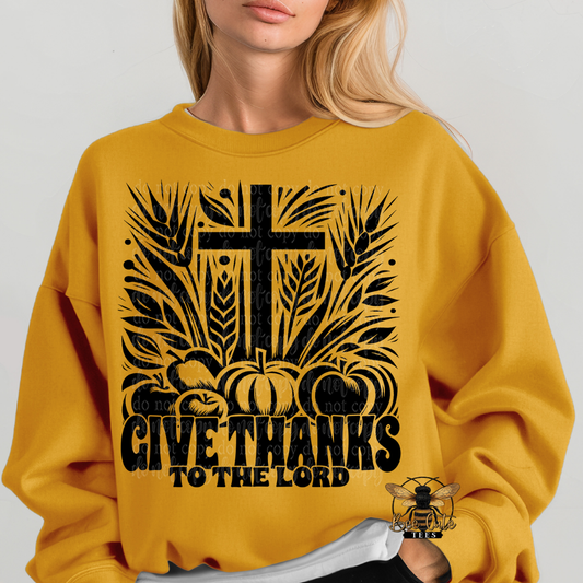 A woman wearing a mustard yellow sweatshirt with a black graphic print featuring a cross, pumpkins, wheat, and the text 'GIVE THANKS TO THE LORD'.
