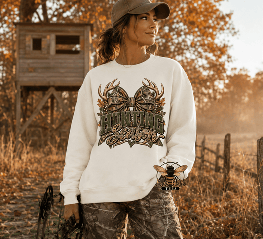 A woman wearing a white crewneck sweatshirt with a camouflage hunting graphic that reads 'HUNTING Season' with antlers and a bow, standing outdoors in a wooded setting with a wooden hunting stand in the background.
