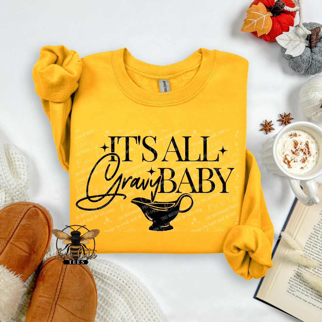 A folded yellow long-sleeve sweatshirt with rolled cuffs, featuring the black text 'IT'S ALL GRAVY BABY' and a black gravy boat graphic on the front. The sweatshirt is styled in a cozy flat-lay arrangement with brown slippers, a mug of hot cocoa, an open book, and autumn decorations on a white background.