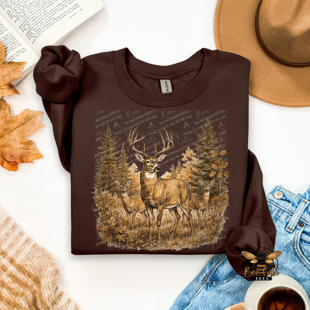 A dark brown long-sleeve crewneck sweatshirt with a graphic of three deer in a forest, styled in a flat lay with a tan hat, blue jeans, a book, autumn leaves, and a cup of coffee.