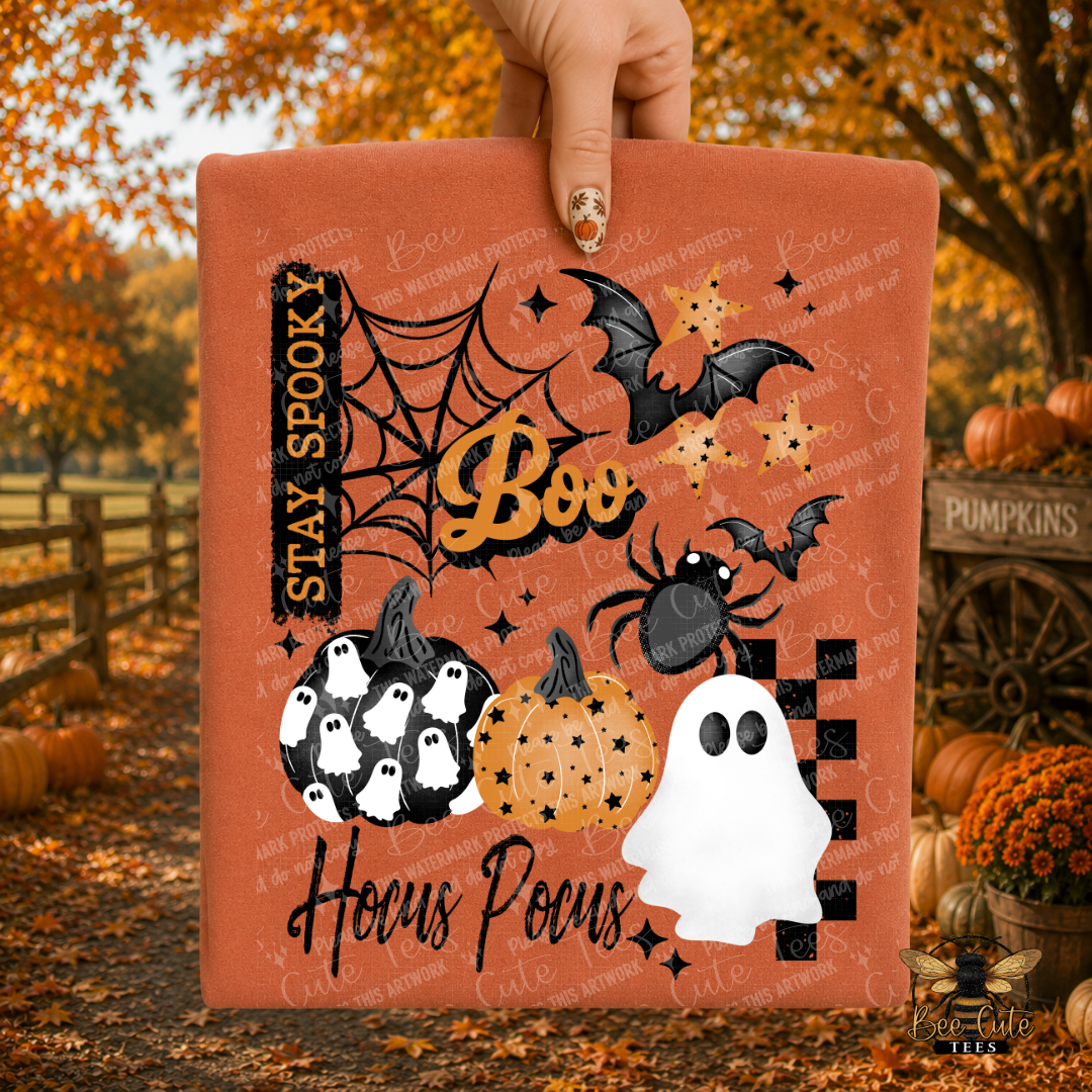 A hand holds a folded orange Halloween-themed t-shirt with printed graphics including bats, a spider, pumpkins, ghosts, stars, and the words “Stay Spooky,” “Boo,” and “Hocus Pocus.” The background shows an autumn outdoor scene with fallen leaves, pumpkins, and a wooden fence.
