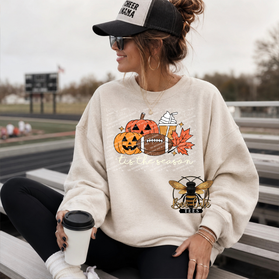 A woman sitting on outdoor stadium bleachers wearing a cream sweatshirt with a fall sports graphic featuring pumpkins, a football, a drink, an autumn leaf, and a bee logo. She is wearing a black and white trucker hat and holding a coffee cup.