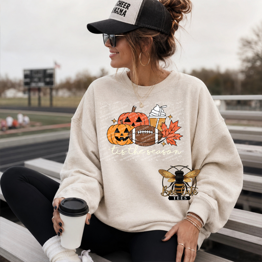 A woman sitting on outdoor stadium bleachers wearing a cream sweatshirt with a fall sports graphic featuring pumpkins, a football, a drink, an autumn leaf, and a bee logo. She is wearing a black and white trucker hat and holding a coffee cup.