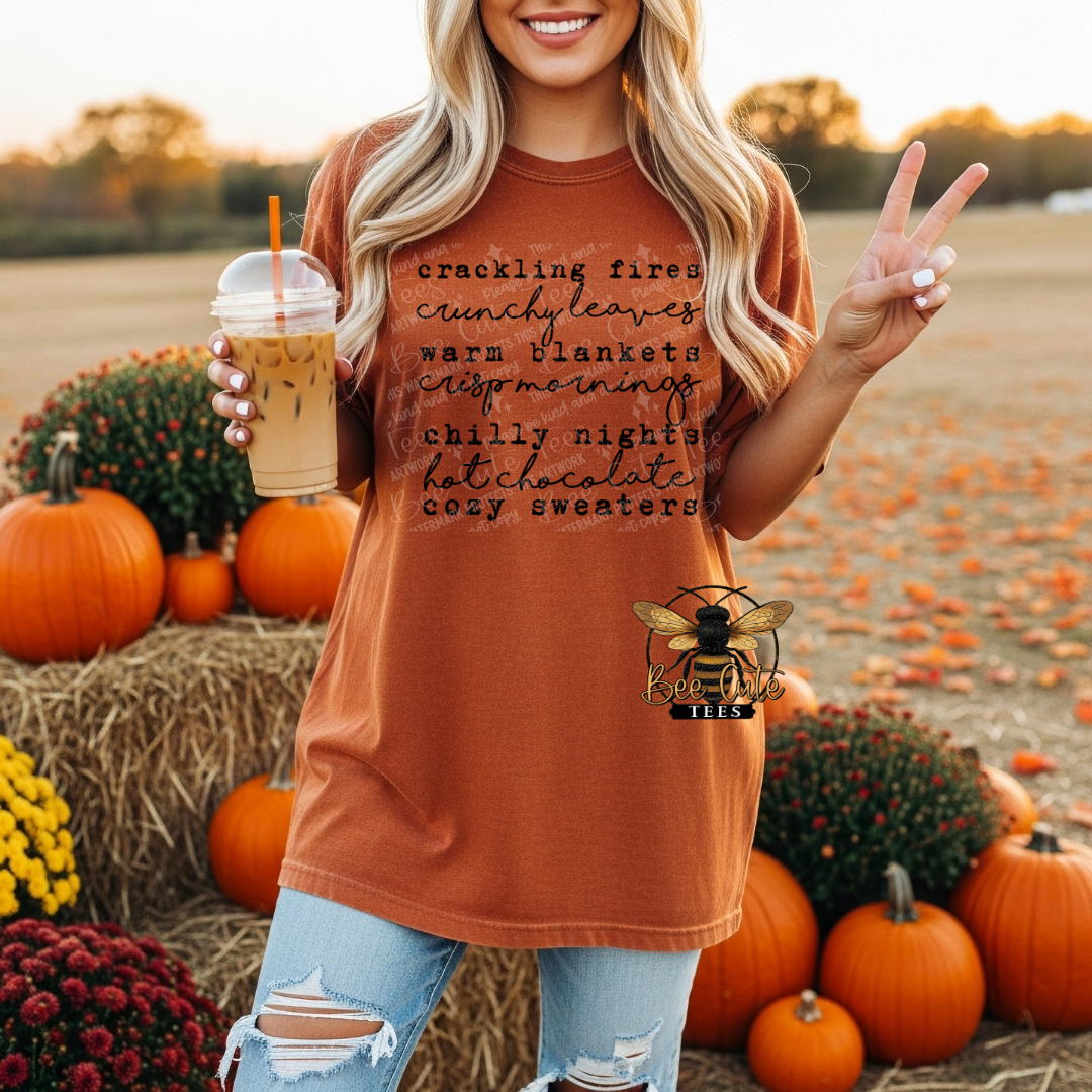 A woman in a rust-orange graphic t-shirt stands in a fall harvest field surrounded by pumpkins and mums, holding an iced coffee. The t-shirt features black script text listing cozy autumn themes.