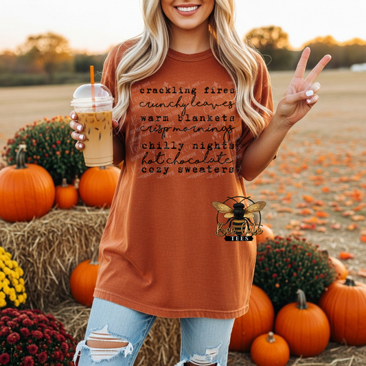 A woman in a rust-orange graphic t-shirt stands in a fall harvest field surrounded by pumpkins and mums, holding an iced coffee. The t-shirt features black script text listing cozy autumn themes.