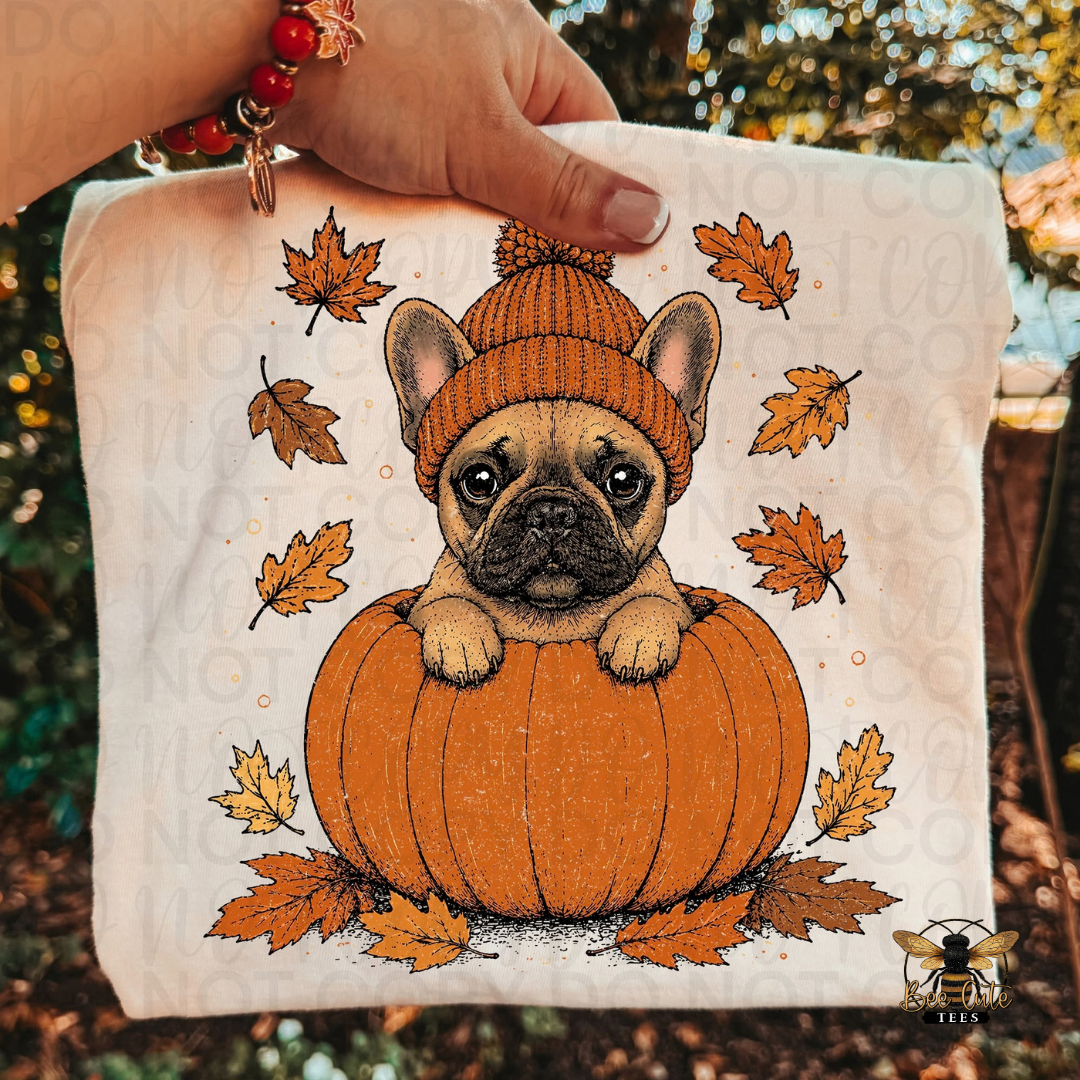 A hand holds a folded beige t-shirt, printed with an illustration of a French bulldog sitting in a pumpkin and surrounded by autumn leaves. The background is a blurred outdoor scene with trees and sunlight.