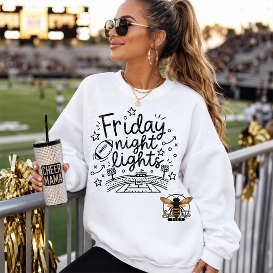 A woman wearing a white graphic sweatshirt with 'Friday Night Lights' and a bee logo, sitting at a stadium railing and holding a glittery tumbler.
