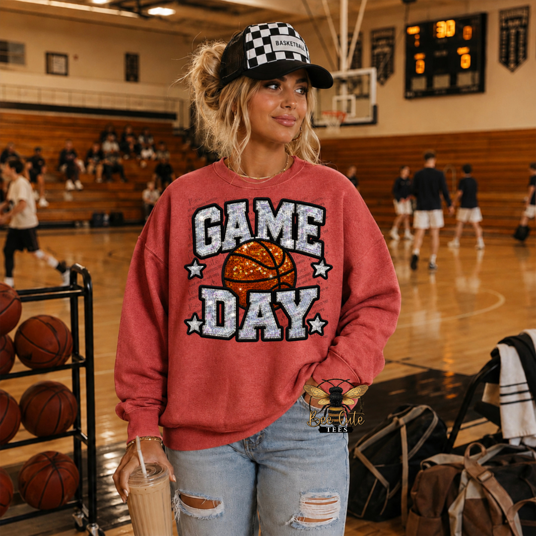 A woman standing on a basketball court wearing a red sweatshirt with a 'GAME DAY' graphic featuring a basketball and distressed white text. She is also wearing a black and white checkered baseball cap and ripped light-wash jeans, holding a drink.
