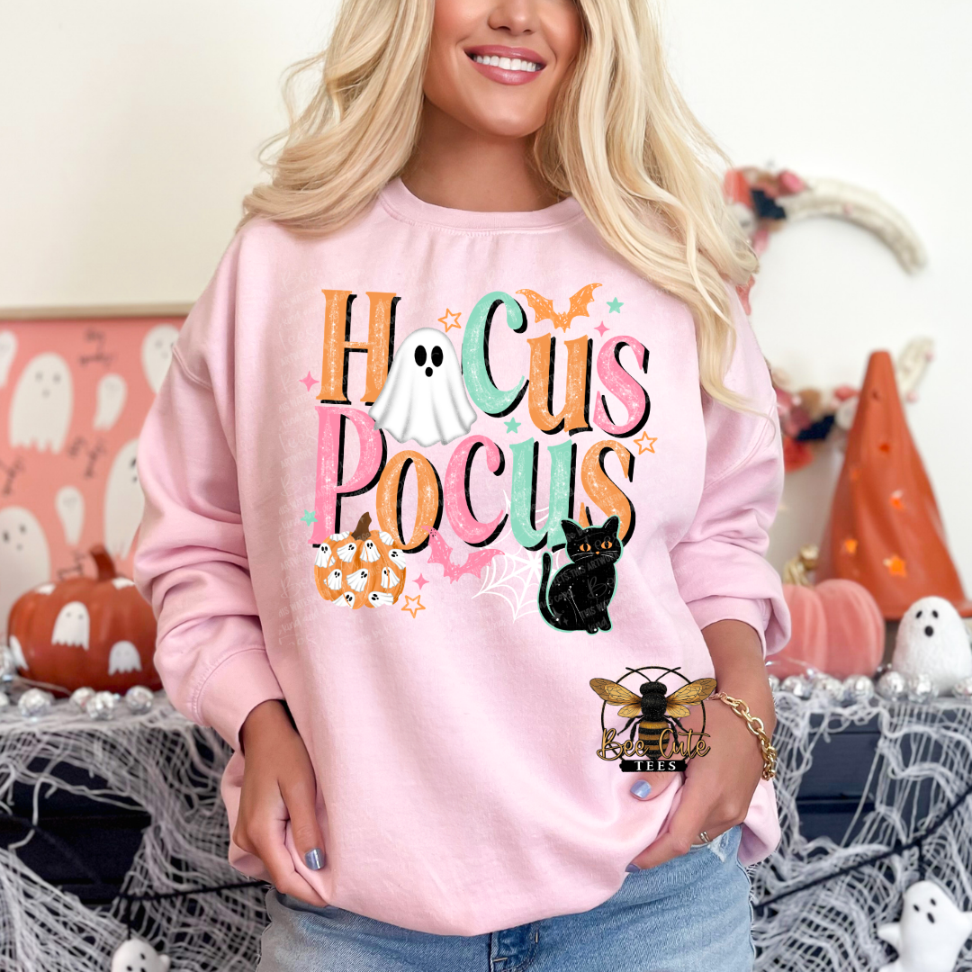 A woman wearing a light pink crewneck sweatshirt with a colorful 'Hocus Pocus' graphic featuring ghosts, a black cat, a spider, and a bee logo. The background is a cozy, decorated Halloween scene with pumpkins, cobwebs, and a ghost figure.