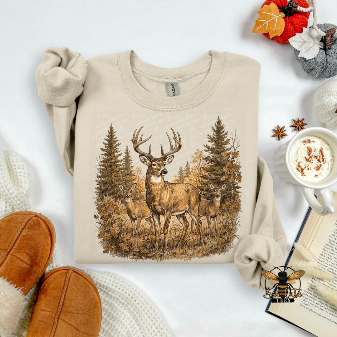 A beige crewneck sweatshirt with a detailed graphic of three deer in a forest, styled in a cozy flat-lay arrangement with a mug of coffee, an open book, and brown slippers.