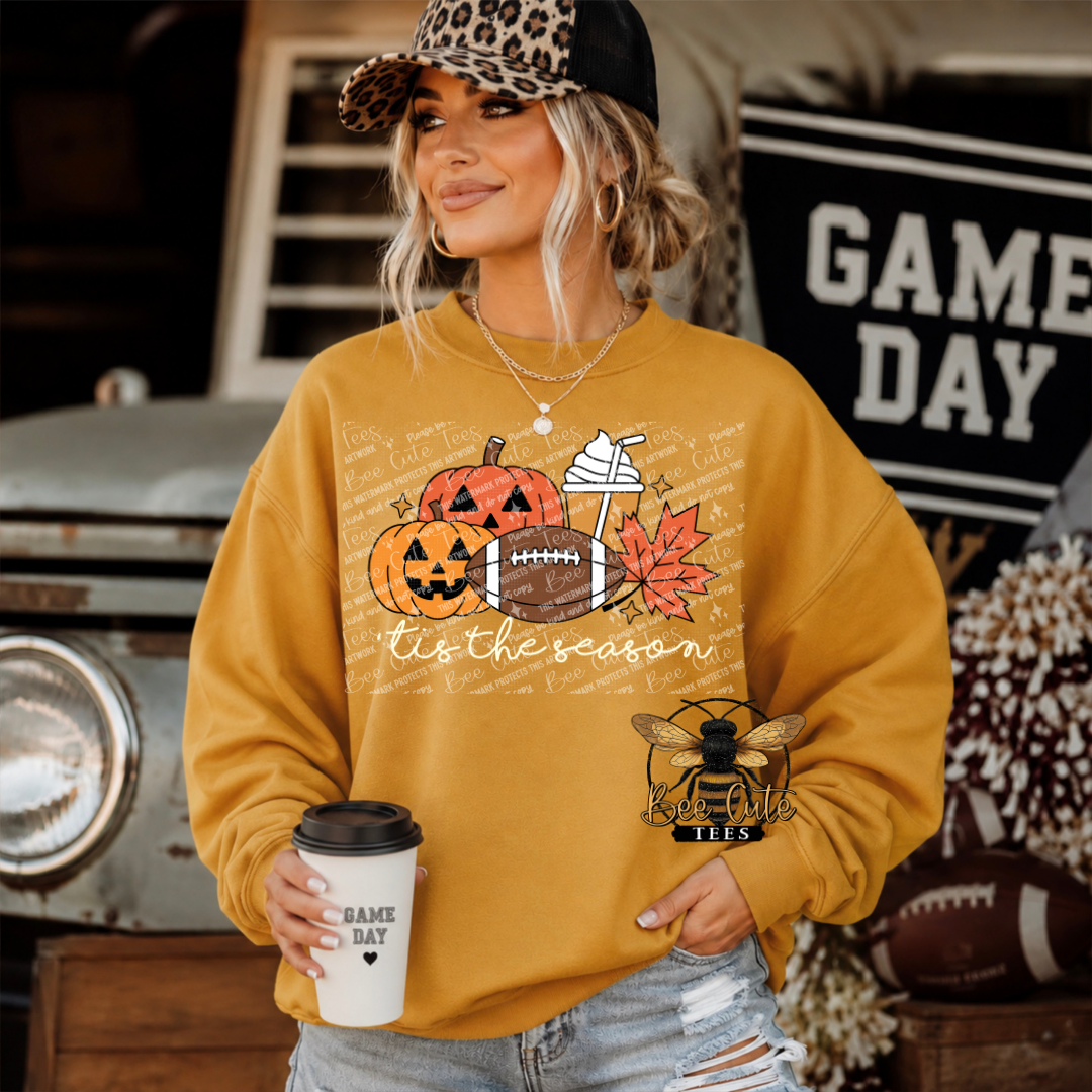 A woman wearing a mustard yellow graphic sweatshirt with a fall sports design, holding a coffee cup in a rustic, autumn-themed setting.