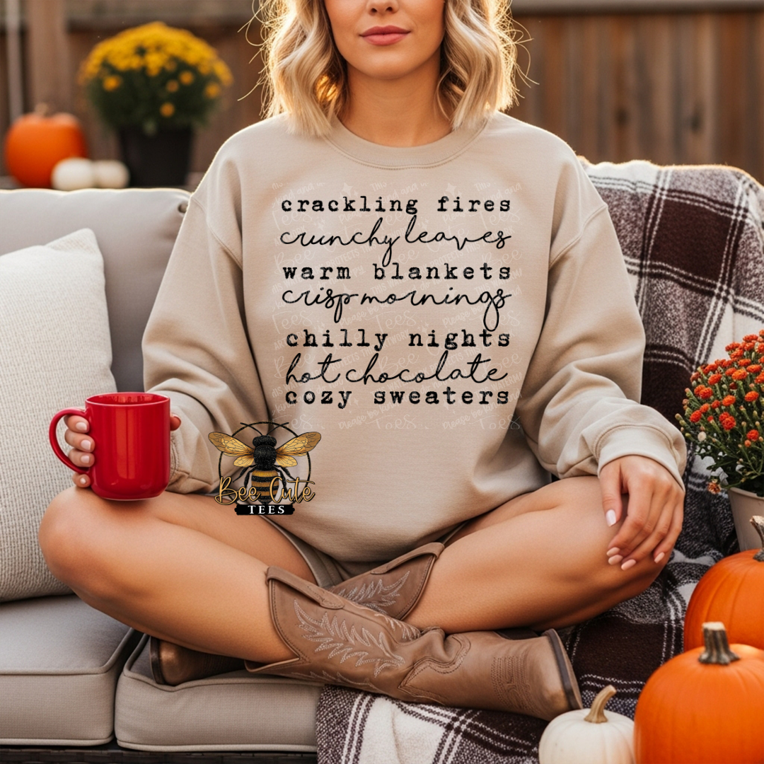 A woman sits cross-legged on a porch wearing a beige crewneck sweatshirt with a black typographic list of cozy autumn phrases. She holds a red mug and wears brown cowboy boots, with pumpkins and a plaid throw nearby.