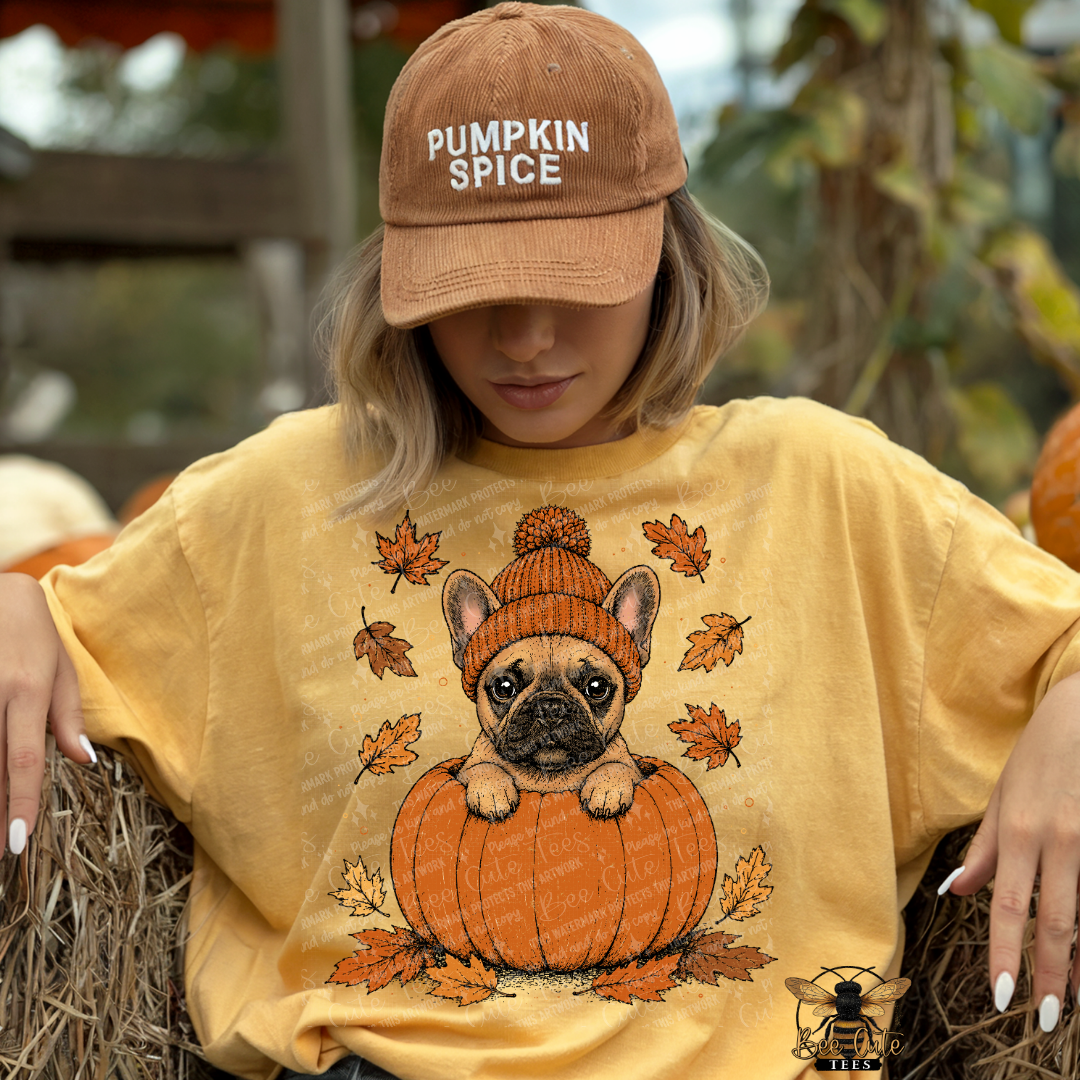 A woman wearing a tan 'Pumpkin Spice' baseball cap and a yellow graphic t-shirt featuring a French Bulldog in a pumpkin, sitting on a hay bale at a pumpkin patch.