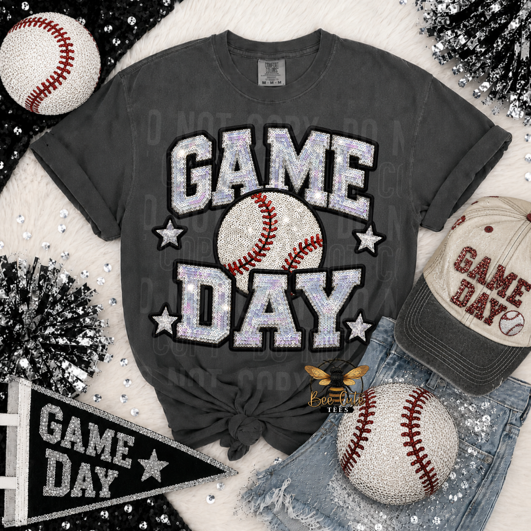 A dark gray graphic t-shirt with 'GAME DAY' in glittery silver letters and a baseball graphic, styled in a flat lay with a baseball cap, denim shorts, and baseballs on a white faux fur background.
