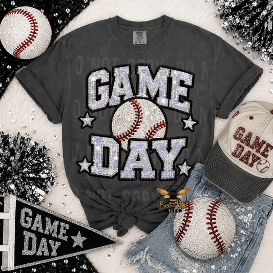 A dark gray graphic t-shirt with 'GAME DAY' in glittery silver letters and a baseball graphic, styled in a flat lay with a baseball cap, denim shorts, and baseballs on a white faux fur background.