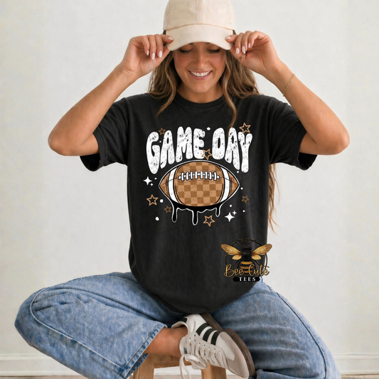 A woman wearing a black graphic t-shirt with a 'Game Day' football design, blue jeans, and a beige cap, sitting on a stool against a light wall.