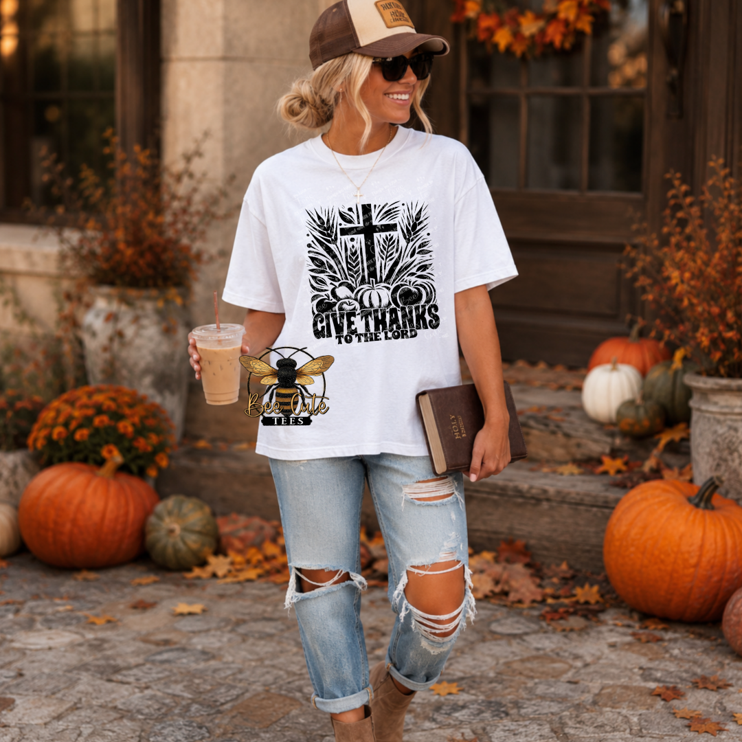 A woman stands outdoors in a fall-themed setting wearing a white graphic t-shirt with a black 'Give Thanks to God' cross design and a bee logo, paired with ripped light-wash jeans and a brown cap.