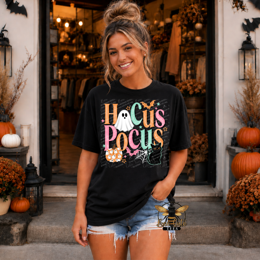 A woman stands in front of a storefront doorway wearing a black graphic T-shirt with a colorful 'Hocus Pocus' Halloween design and denim shorts. The scene is decorated with pumpkins, fall foliage, and lanterns.