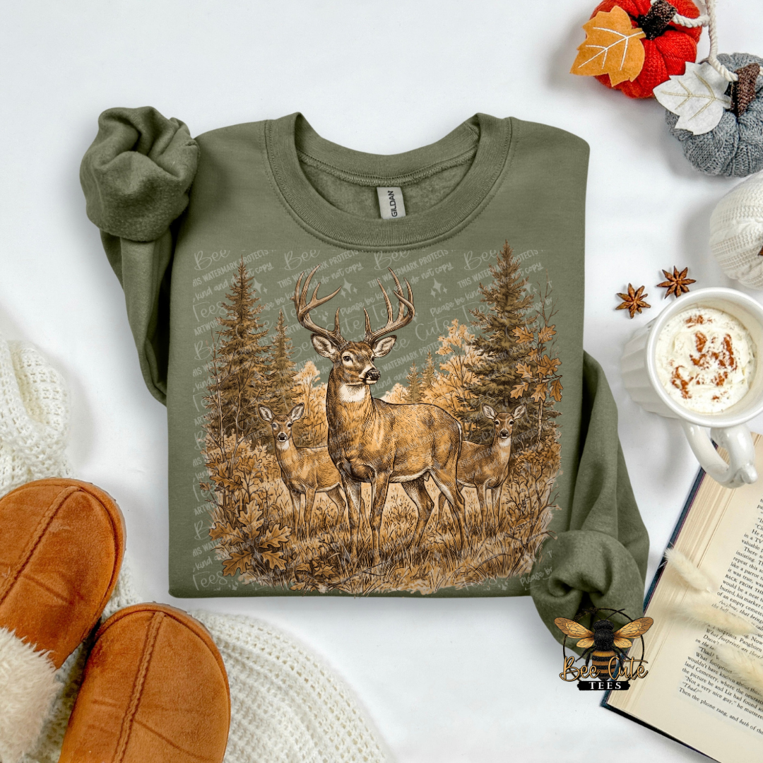 An olive green long-sleeve crewneck sweatshirt with a detailed graphic of three deer in a pine forest, styled in a cozy flat-lay arrangement with a mug of coffee, an open book, and brown slippers.
