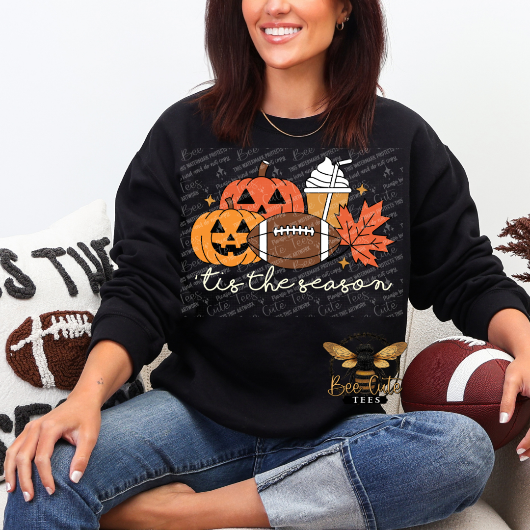 A woman wearing a black crewneck sweatshirt with a festive autumn graphic that reads 'tis the season' and features jack-o'-lanterns, a football, an orange leaf, and a coffee cup. She is sitting on a couch holding a football, with a decorative pillow in the background.
