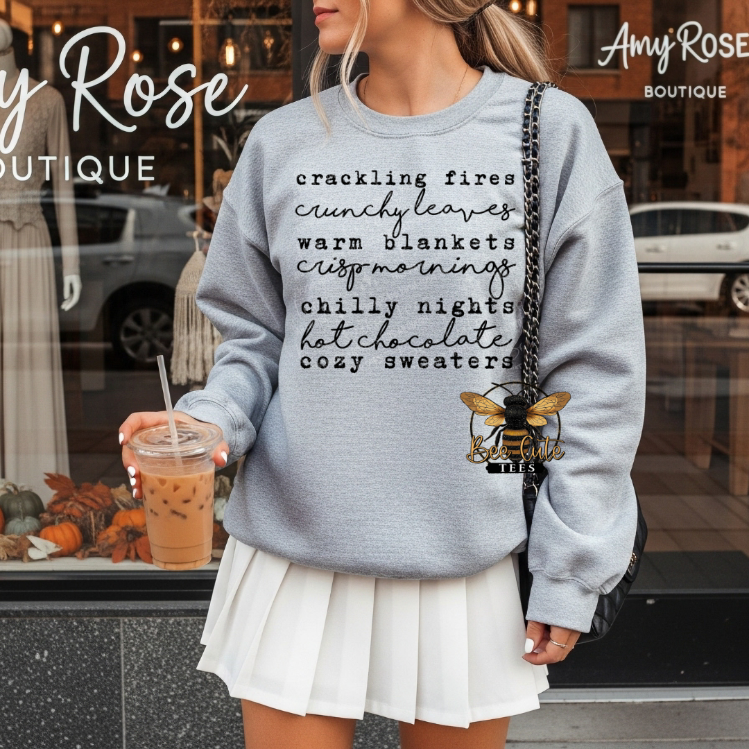 A woman wearing a light gray crewneck sweatshirt with a black typographic print listing cozy autumn phrases, paired with a white pleated skirt and a black shoulder bag. She is holding an iced coffee in front of a boutique storefront.