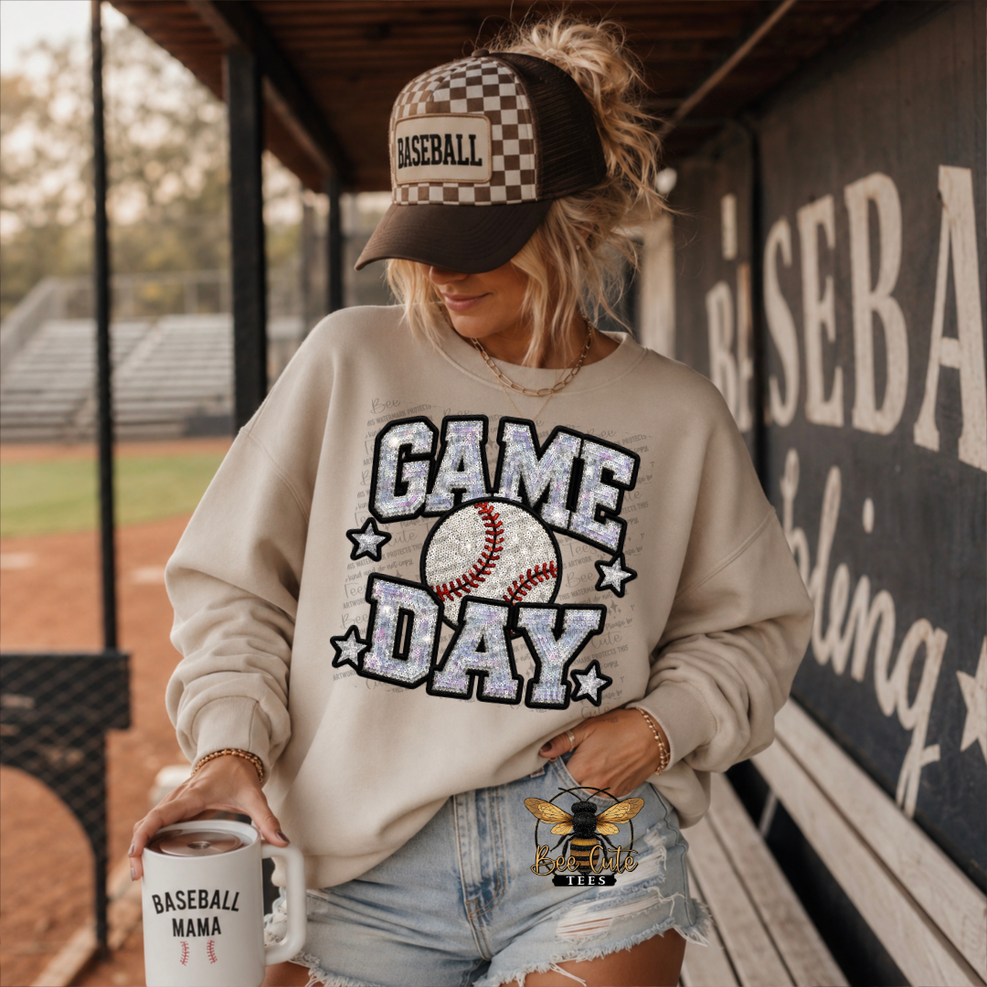 A woman in a baseball dugout wearing a beige sweatshirt with a glittery 'GAME DAY' graphic, a checkered baseball cap, and denim shorts, holding a white mug that says 'BASEBALL MAMA'.