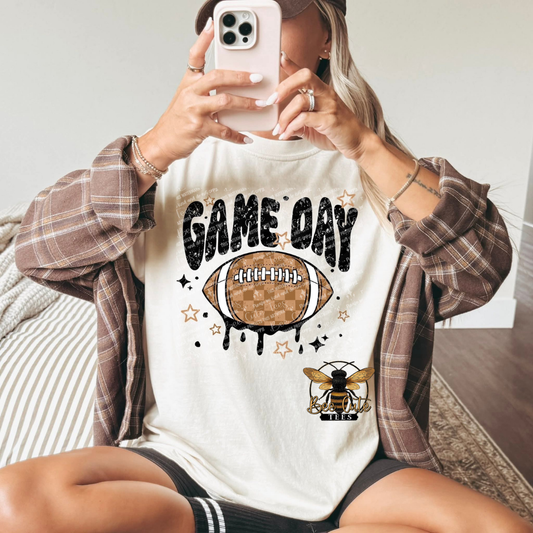 A woman takes a mirror selfie wearing a white graphic t-shirt with a 'GAME DAY' football design, styled with a brown plaid flannel shirt and a brown cap.