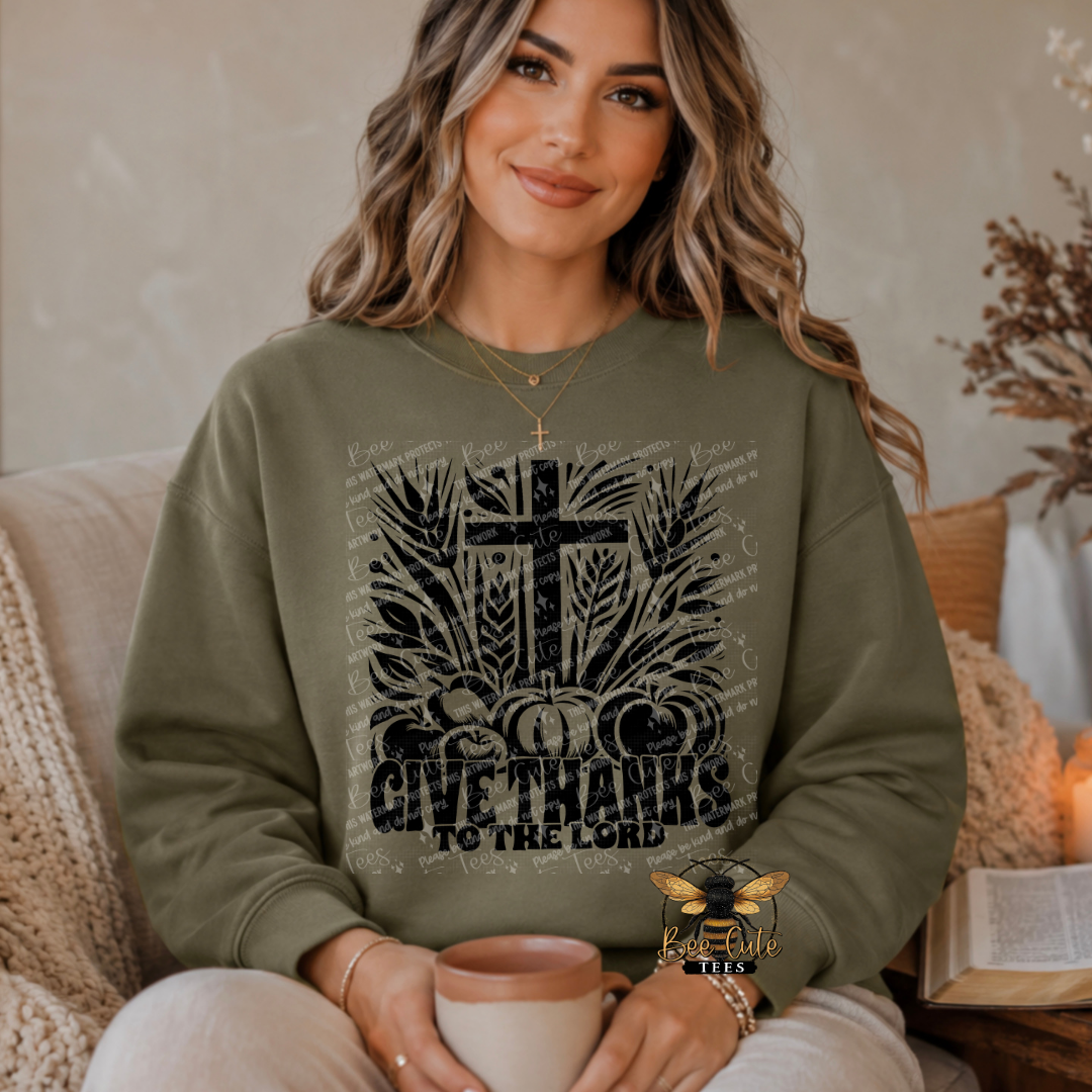 A woman wearing an olive green crewneck sweatshirt with a distressed black graphic of a cross, pumpkins, wheat, and the text 'GIVE THANKS TO THE LORD'. She is holding a mug while sitting on a couch in a cozy indoor setting.
