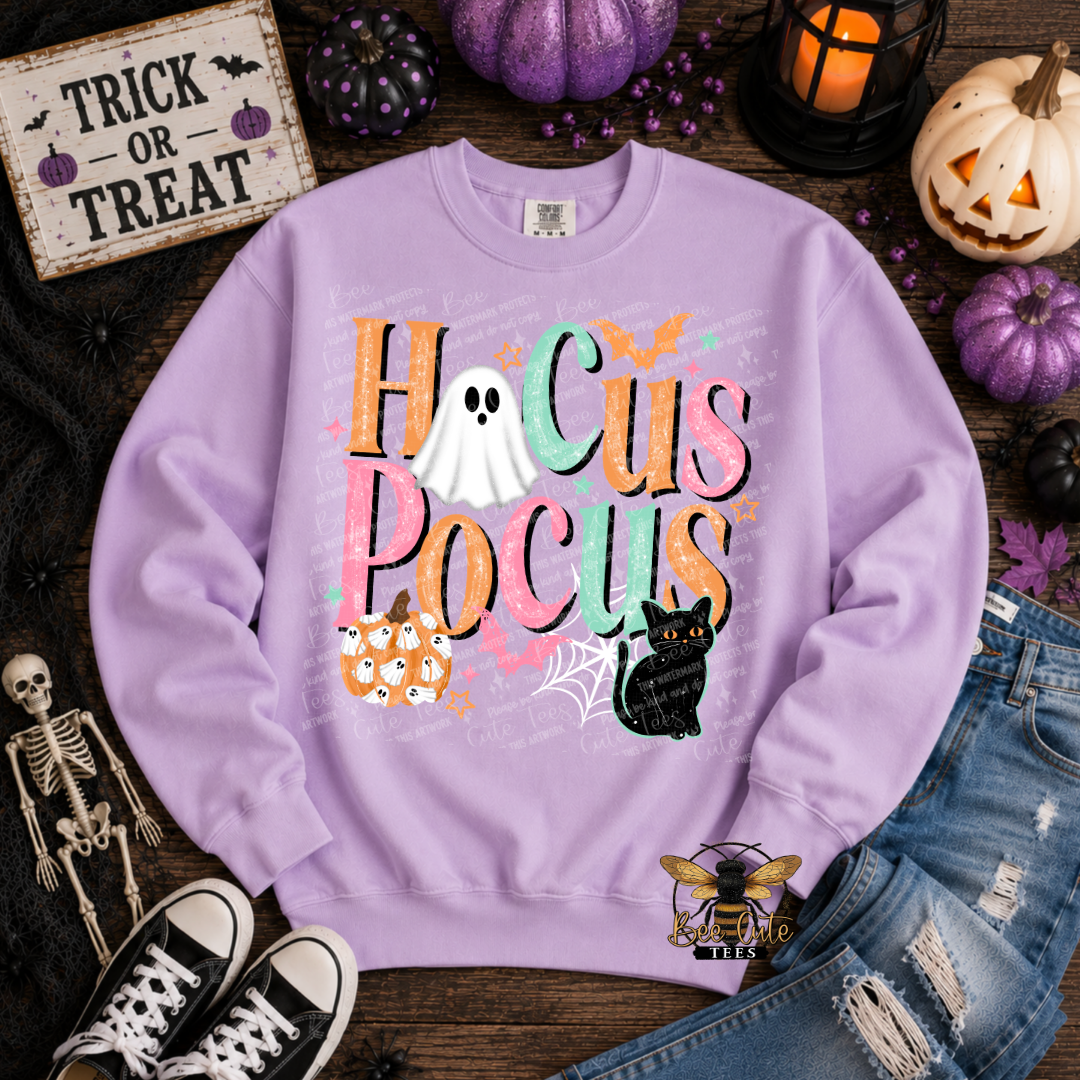 A lavender crewneck sweatshirt with a colorful 'Hocus Pocus' graphic, styled in a flat lay with Halloween props including pumpkins, a skeleton, jeans, and sneakers on a dark wood background.
