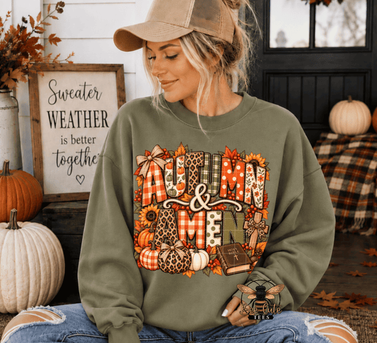 A woman wearing an olive green crewneck sweatshirt with a large 'Autumn & amen' graphic featuring autumn patterns, sitting in a cozy fall-themed room with pumpkins and a framed sign.