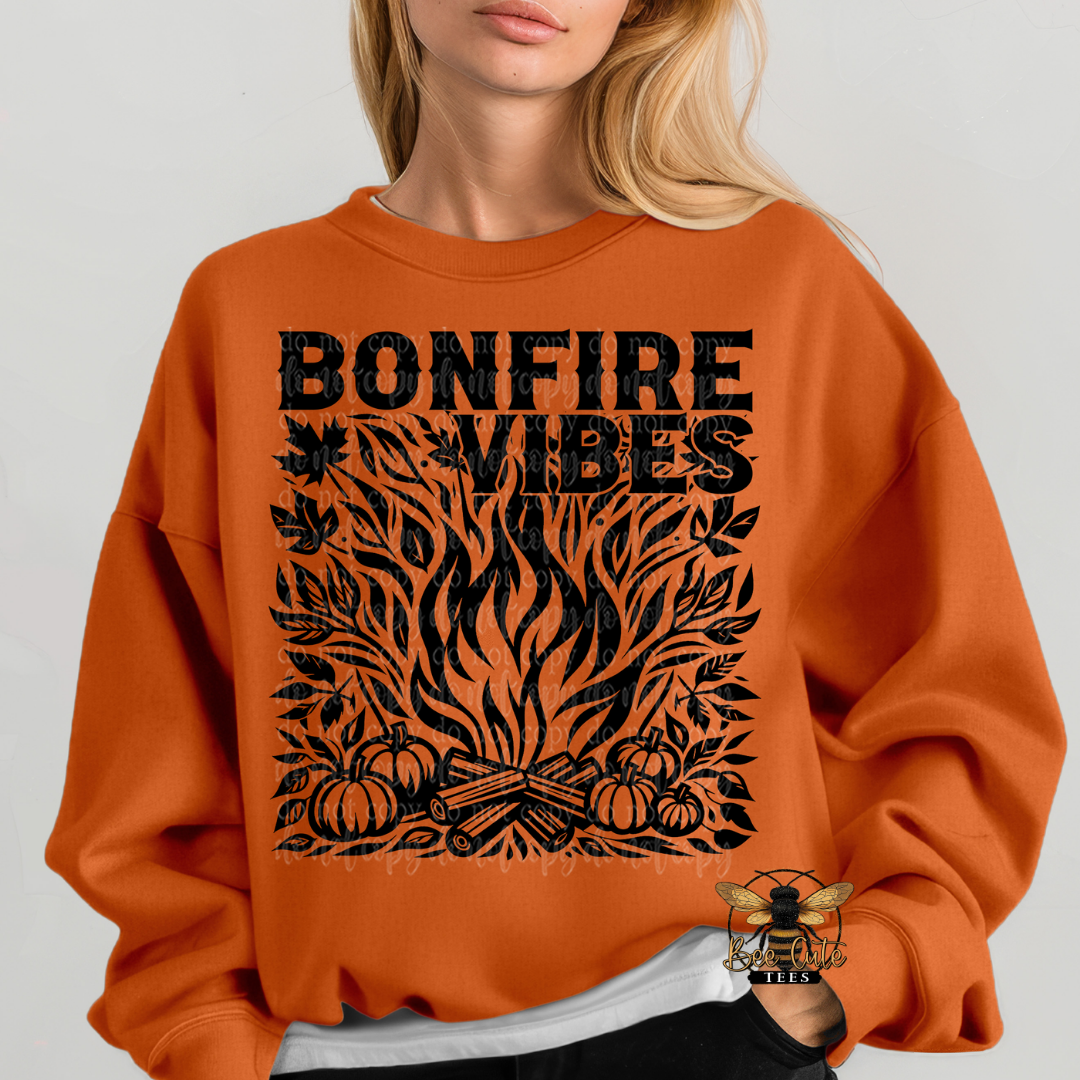A woman wearing a burnt orange crewneck sweatshirt with a black graphic print that reads 'BONFIRE VIBES' above a stylized campfire, pumpkins, and autumn leaves. A bee logo with the text 'BEE CUTE TEES' is overlaid in the bottom right corner.